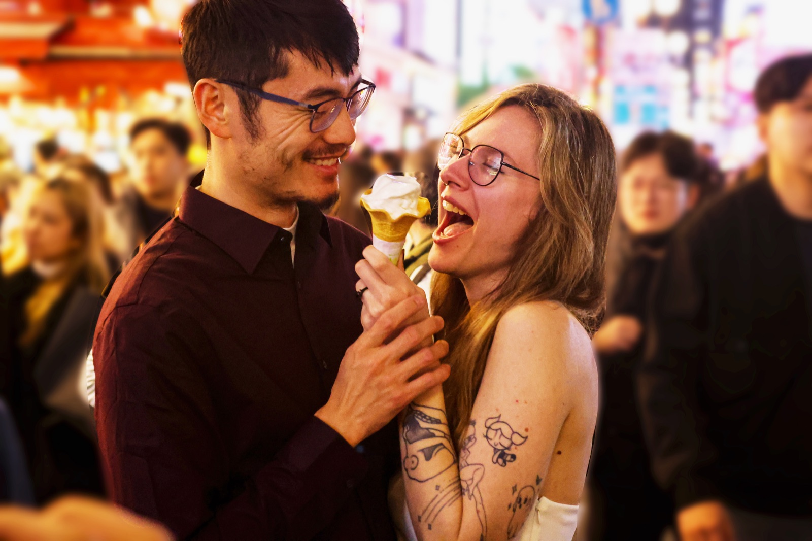 The couple laughing together as one takes a bite of an ice cream cone in a crowded night market.
